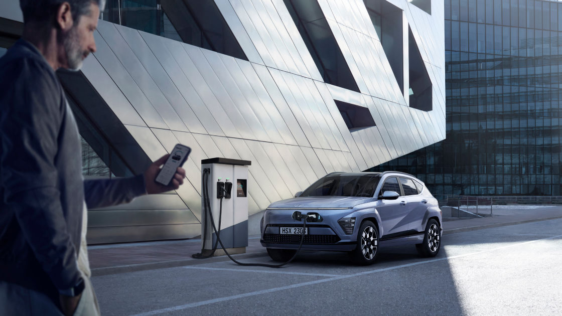 The all-new KONA Electric being charged at the charging station is in the middle of the picture, and a side view of a man checking the condition of The all-new KONA Electric with a smartphone is seen on the left side of the company far away from The all-new KONA Electric.